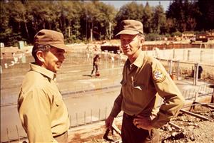 Two men in brown caps and uniform shirts face each other. Behind them are rows of shallow water with trees in the distance. 